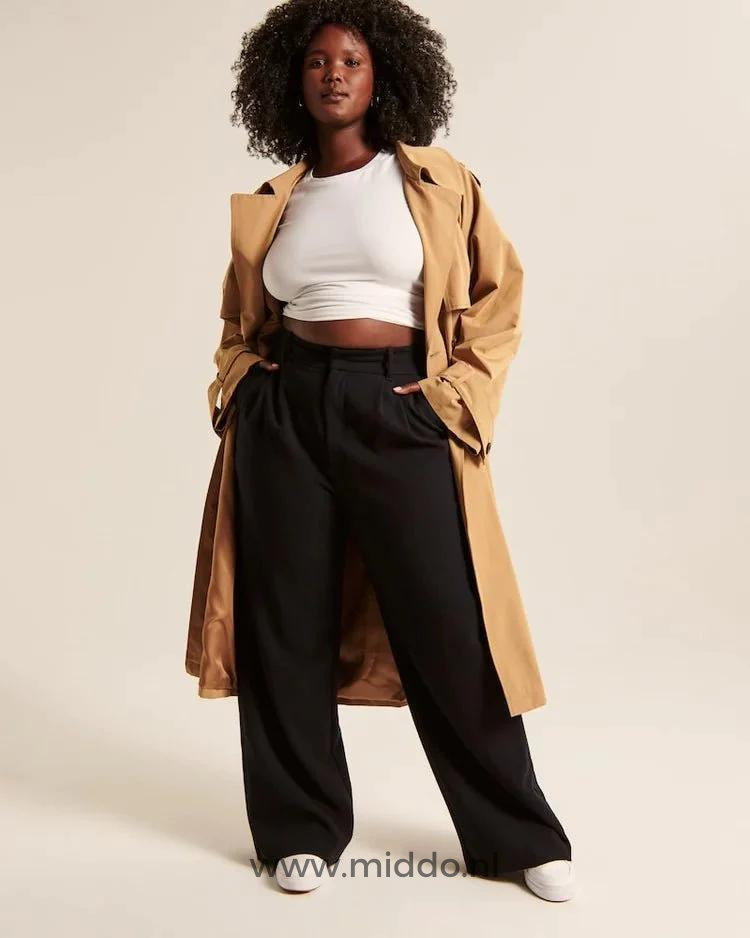 Een donkere getinte dame met lange beige jas showt de zwarte wijde pantalon met witte sportschoenen eronder